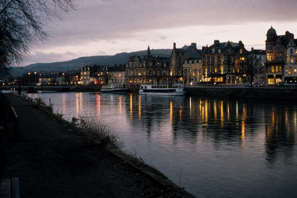 The River Ness through Inverness at dusk, stone bridges and lit windows, hills to the south where the loch begins